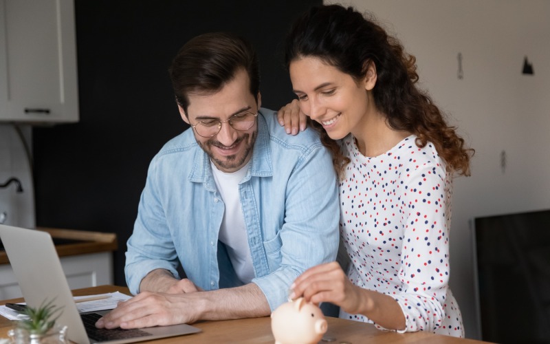 couple in domestic kitchen-with-laptop-putting coin in piggybank